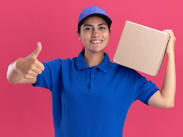 Lächelndes junges Liefermädchen, das Uniform mit Kappe hält Box auf Schulter trägt und Daumen oben auf rosa Wand zeigt