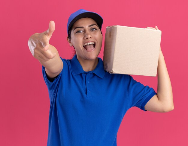 Lächelndes junges Liefermädchen, das Uniform mit Kappe hält Box auf Schulter trägt, zeigt Sie Geste lokalisiert auf rosa Wand