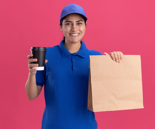 Lächelndes junges Liefermädchen, das Uniform mit Kappe hält, die Papiernahrungsmittelpaket mit Tasse Kaffee lokalisiert auf rosa Wand hält