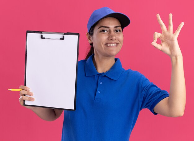 Lächelndes junges Liefermädchen, das Uniform mit Kappe hält Zwischenablage hält und okay Geste auf rosa Wand isoliert zeigt