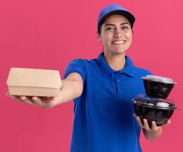 Lächelndes junges Liefermädchen, das Uniform mit Kappe trägt, die Lebensmittelbehälter an der Front lokalisiert auf rosa Wand hält