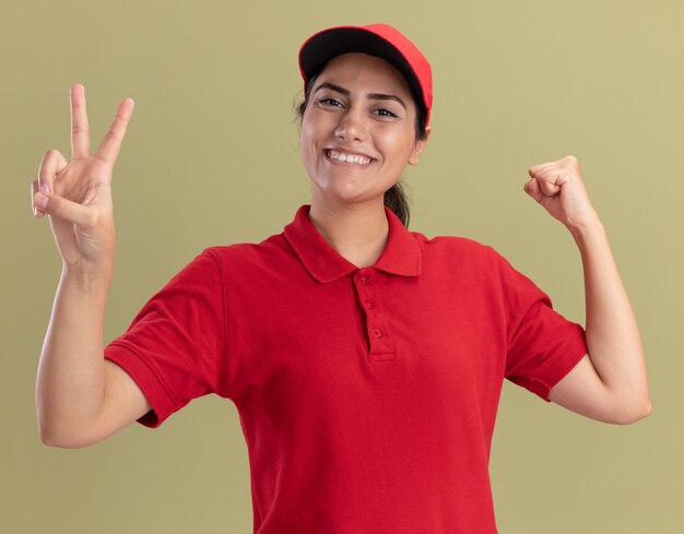 Lächelndes junges Liefermädchen, das Uniform mit Kappe zeigt, die Friedensgeste zeigt, die starke Geste tut, die auf olivgrüner Wand lokalisiert wird