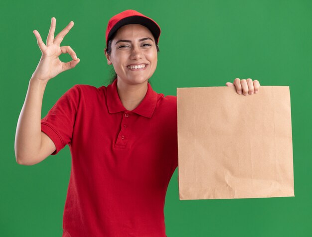 Lächelndes junges Liefermädchen, das Uniform und Kappe hält, die Papiernahrungsmittelpaket hält, das okay Geste lokalisiert auf grüner Wand zeigt