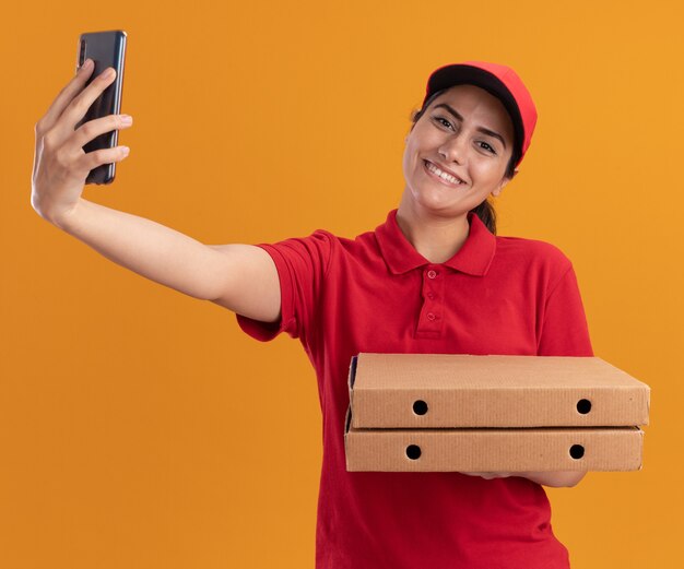 Lächelndes junges Liefermädchen, das Uniform und Kappe hält, die Pizzaschachteln halten und ein Selfie lokalisiert auf orange Wand nehmen