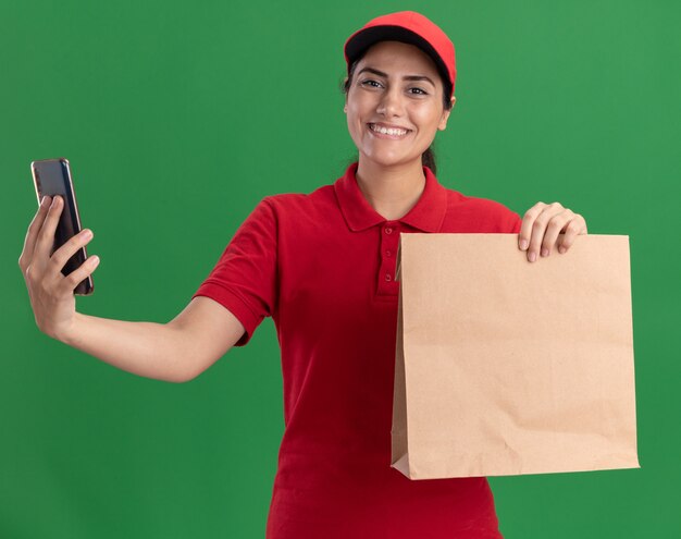 Lächelndes junges Liefermädchen, das Uniform und Kappe trägt, die Papiernahrungsmittelpaket mit Telefon lokalisiert auf grüner Wand hält