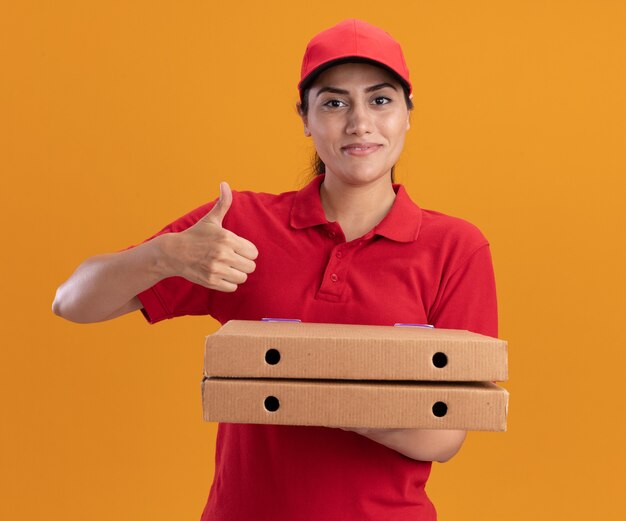 Lächelndes junges Liefermädchen, das Uniform und Kappe trägt, die Pizzakästen hält, die Daumen oben auf orange Wand lokalisiert zeigen