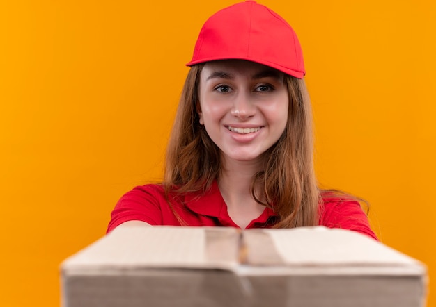 Kostenloses Foto lächelndes junges liefermädchen in der roten uniform, die kasten auf isolierter orange wand ausdehnt