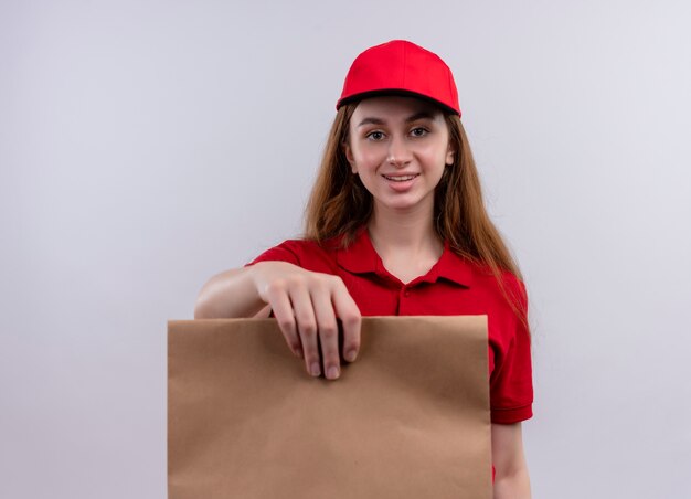 Lächelndes junges Liefermädchen in der roten Uniform, die Papiertüte auf isolierter weißer Wand ausdehnt