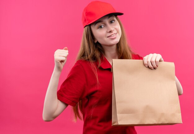 Lächelndes junges Liefermädchen in der roten Uniform, die Papiertüte mit geballter Faust auf isolierter rosa Wand hält