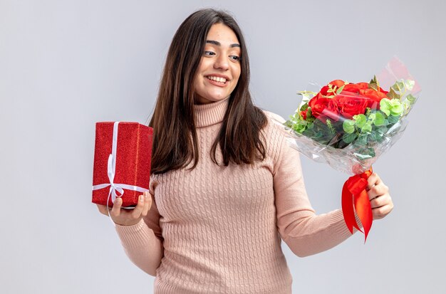 Lächelndes junges Mädchen am Valentinstag mit Geschenkbox und Blick auf Blumenstrauß in der Hand isoliert auf weißem Hintergrund