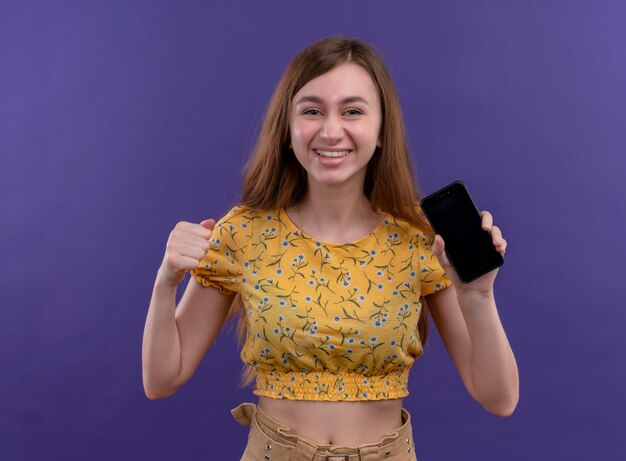 Lächelndes junges Mädchen, das Handy hält und Faust auf isolierter lila Wand erhebt