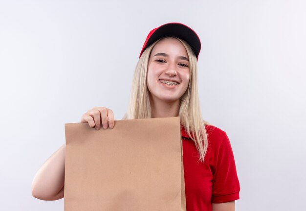 Lächelndes junges Mädchen der Lieferung, das rotes T-Shirt und Kappe in der Zahnspange trägt, die Papiertasche auf lokalisiertem weißem Hintergrund hält