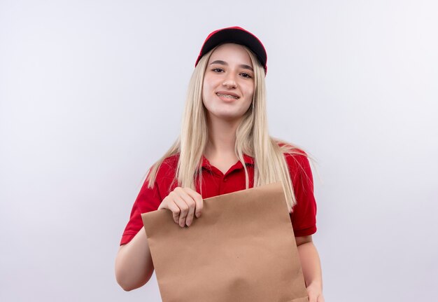 Lächelndes junges Mädchen der Lieferung, das rotes T-Shirt und Kappe in der Zahnspange trägt, die Papiertasche auf lokalisiertem weißem Hintergrund hält