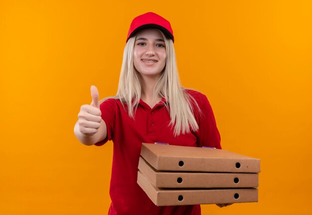 Lächelndes junges Mädchen der Lieferung, das rotes T-Shirt und Kappe in der Zahnspange trägt, die Pizza bax ihren Daumen oben auf lokalisiertem orangefarbenem Hintergrund hält