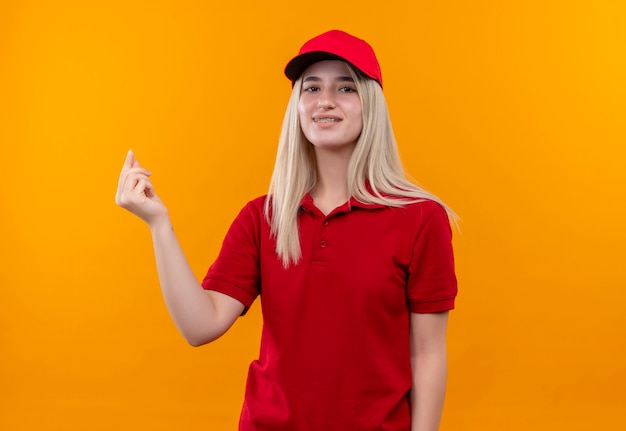 Lächelndes junges Mädchen der Lieferung, das rotes T-Shirt und Kappe in der Zahnspange trägt, die Spitzengeste auf lokalisiertem orange Hintergrund zeigt