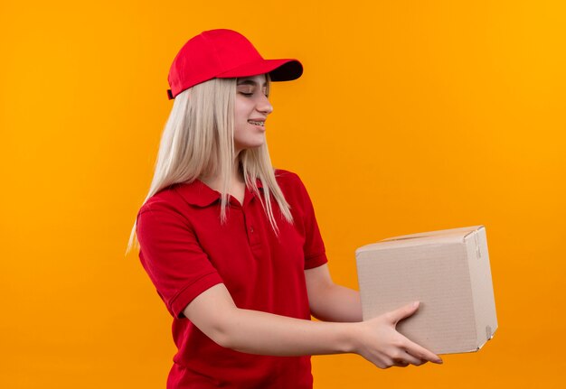 Lächelndes junges Mädchen der Lieferung, das rotes T-Shirt und Kappe in der Zahnspangenhaltebox an der Seite auf lokalisiertem orange Hintergrund trägt