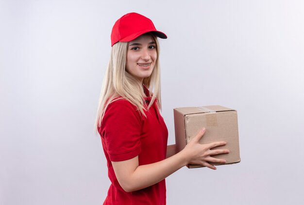 Lächelndes junges Mädchen der Lieferung, das rotes T-Shirt und Kappe in der Zahnspangenhaltebox auf lokalem weißem Hintergrund trägt