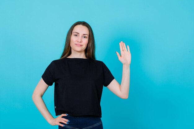 Lächelndes junges Mädchen hebt eine Hand und legt die andere auf die Taille auf blauem Hintergrund