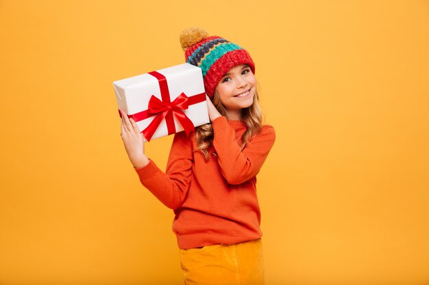 Lächelndes junges Mädchen in der Strickjacke und in Hut, die Geschenkbox halten und die Kamera über Orange betrachten