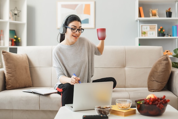 Lächelndes junges Mädchen mit Brille und Kopfhörern, das eine Tasse Tee auf dem Sofa hinter dem Couchtisch mit Blick auf den Laptop im Wohnzimmer hält