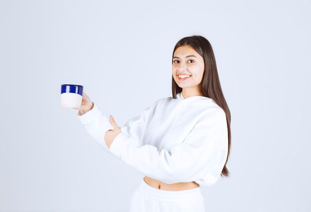 lächelndes junges Mädchen mit einer Tasse und zeigt einen Daumen nach oben auf weiß-grauem Hintergrund.