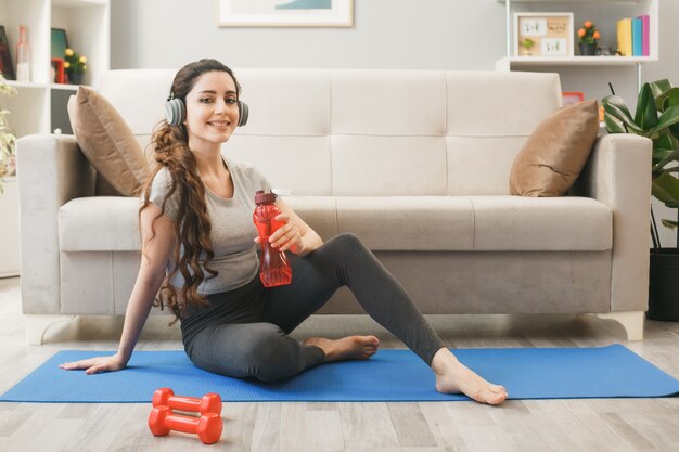 Lächelndes junges Mädchen mit Kopfhörern, das eine Wasserflasche vor dem Sofa im Wohnzimmer hält?