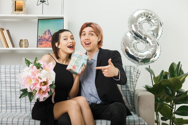 Lächelndes junges Paar am glücklichen Frauentag hält und zeigt auf Blumenstrauß und Gegenwart, die auf dem Sofa im Wohnzimmer sitzt