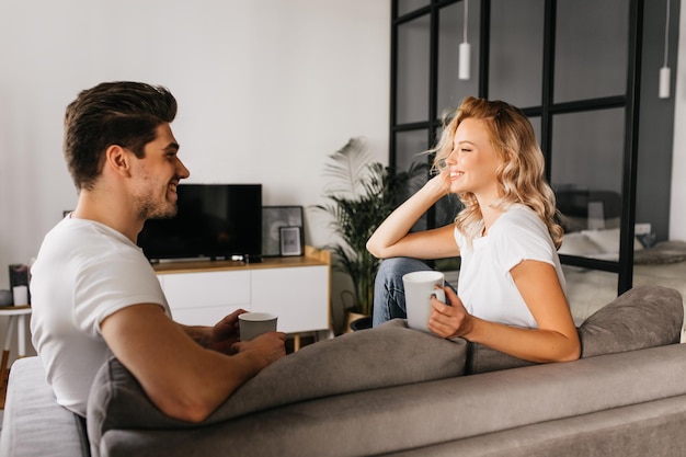 Lächelndes junges Paar, das zu Hause sitzt und einander ansieht und Tassen hält Gemütliches Heimfoto von zwei attraktiven verliebten Menschen, die Zeit miteinander verbringen