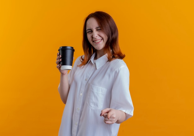 Lächelndes junges rothaariges Mädchen, das Tasse Kaffee hält und Sie Geste lokalisiert auf gelber Wand zeigt