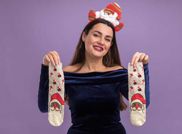 Lächelndes junges schönes Mädchen, das blaues Kleid und Weihnachtshaarbügel hält Weihnachtssocken lokalisiert auf lila Hintergrund trägt