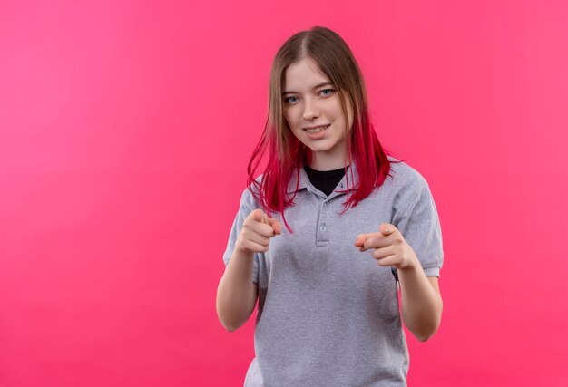 Lächelndes junges schönes Mädchen, das graues T-Shirt trägt, das Sie Geste auf lokalisiertem rosa Hintergrund zeigt