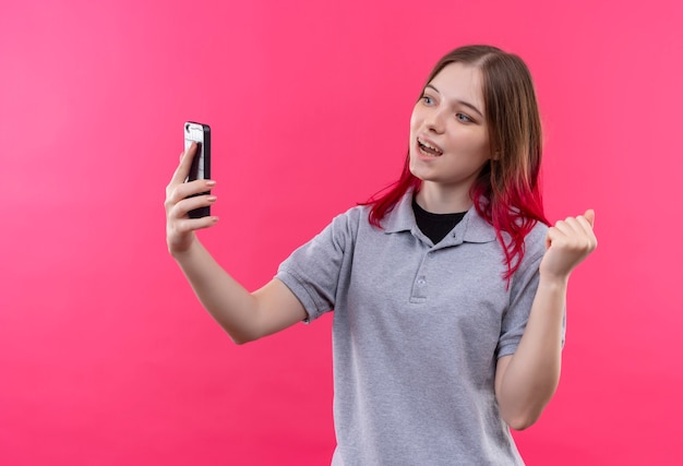 Lächelndes junges schönes Mädchen, das graues T-Shirt trägt, das Telefon in ihrer Hand betrachtet, die ja Geste auf lokalisiertem rosa Hintergrund zeigt