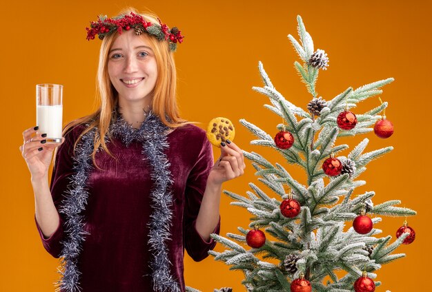 lächelndes junges schönes Mädchen, das in der Nähe des Weihnachtsbaums steht und ein rotes Kleid und einen Kranz mit Girlande am Hals trägt, die ein Glas Milch mit Keksen isoliert auf oranger Wand hält