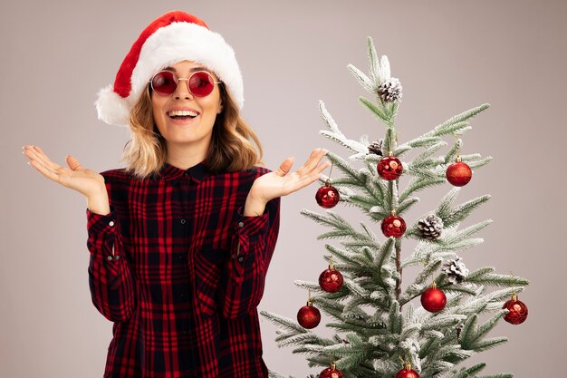 Lächelndes junges schönes Mädchen, das in der Nähe des Weihnachtsbaums steht und Weihnachtsmütze mit Brille trägt, die die Hände auf weißem Hintergrund ausbreitet