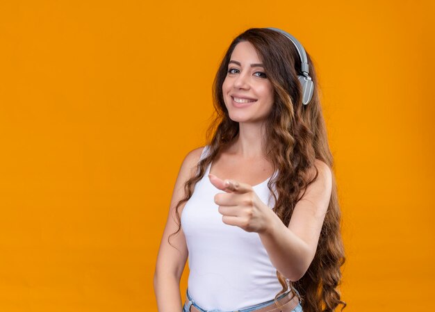 Lächelndes junges schönes Mädchen, das Kopfhörer trägt, die auf isolierten orange Raum mit Kopienraum zeigen