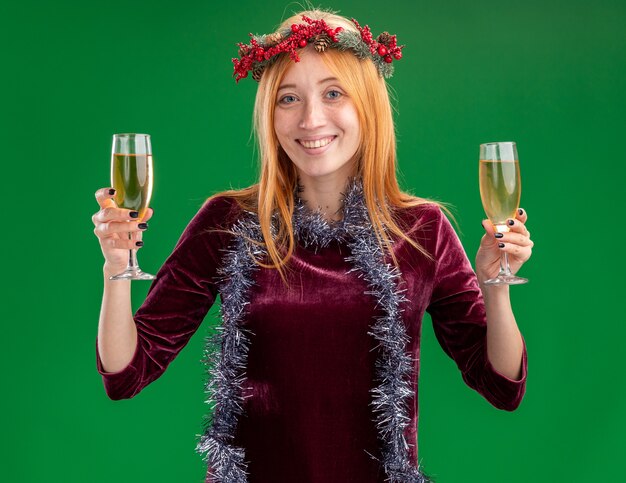 Lächelndes junges schönes Mädchen, das rotes Kleid mit Kranz und Girlande am Hals hält Glas des Champagners lokalisiert auf grünem Hintergrund trägt