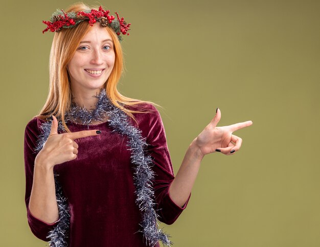 Lächelndes junges schönes Mädchen, das rotes Kleid mit Kranz und Girlande auf Halspunkten an der Seite lokalisiert auf olivgrünem Hintergrund mit Kopienraum trägt