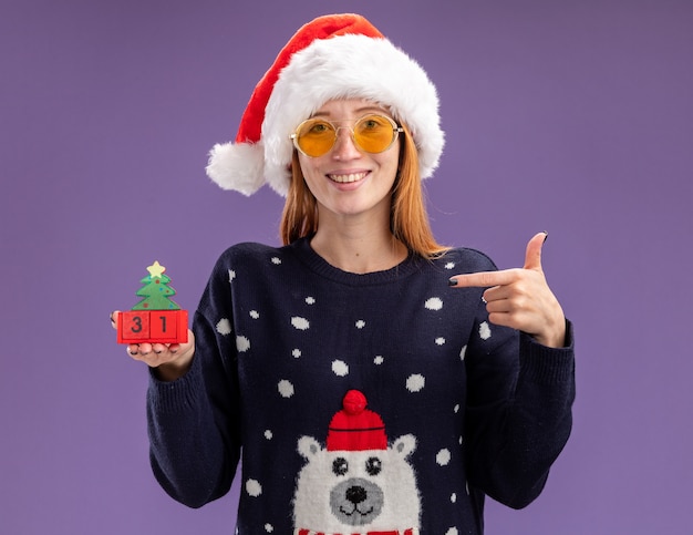 Lächelndes junges schönes Mädchen, das Weihnachtspullover und Hut mit Brille hält und Punkte auf Weihnachtsspielzeug lokalisiert auf lila Hintergrund trägt