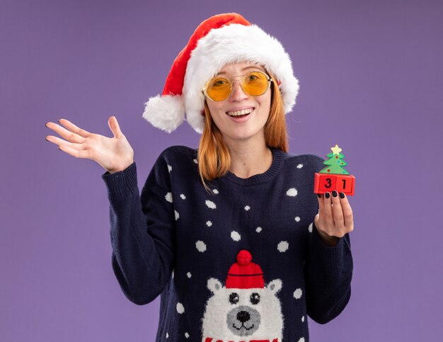 Lächelndes junges schönes Mädchen, das Weihnachtspullover und Hut mit Gläsern hält, die Weihnachtsspielzeug spreizende Hände lokalisiert auf lila Hintergrund tragen