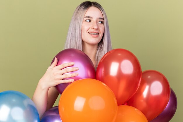 Lächelndes junges schönes Mädchen, das Zahnspangen trägt, die hinter Ballons stehen, die auf olivgrüner Wand isoliert sind?