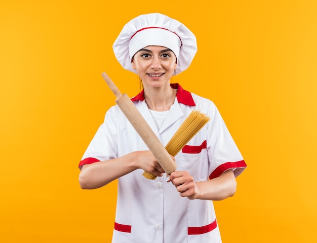 Lächelndes junges schönes Mädchen in Kochuniform, das Spaghetti mit Nudelholz hält und überquert