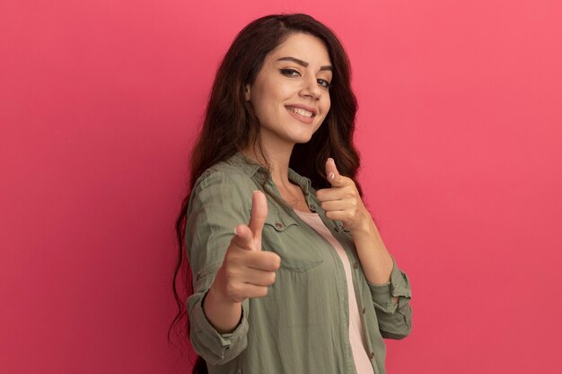 Lächelndes junges schönes Mädchen mit olivgrünem T-Shirt zeigt auf die Kamera isoliert auf rosa Wand