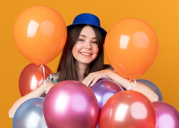 Lächelndes junges schönes Mädchen mit Partyhut, das hinter Luftballons steht
