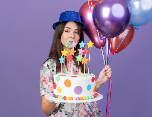 Lächelndes junges schönes Mädchen mit Partyhut, das Luftballons mit Kuchen hält, isoliert auf blauer Wand