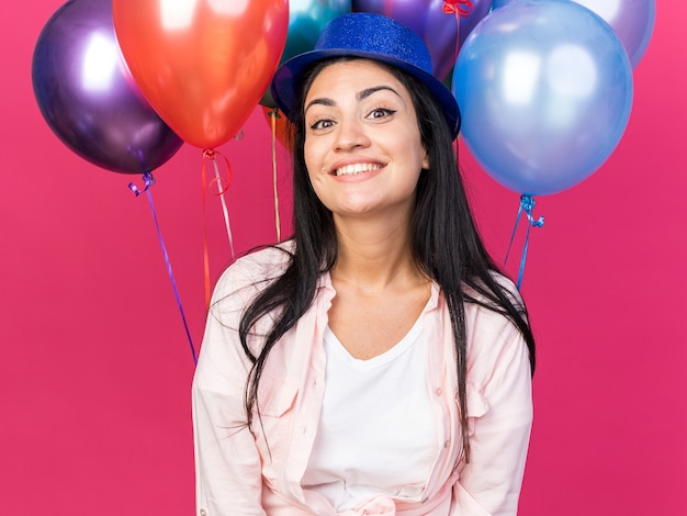 Lächelndes junges schönes Mädchen mit Partyhut, das vor Ballons steht