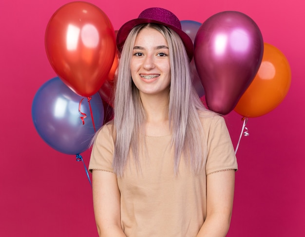 Lächelndes junges schönes Mädchen mit Partyhut mit Hosenträgern, die vor Ballons stehen