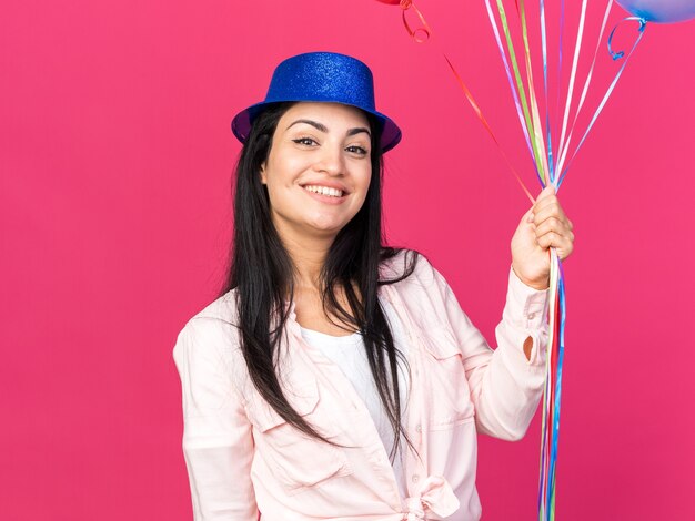 Lächelndes junges schönes Mädchen mit Partyhut mit Luftballons isoliert auf rosa Wand