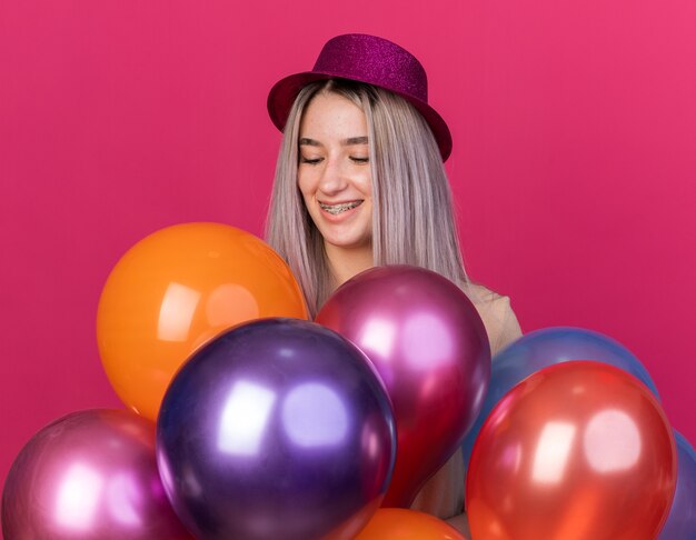 Lächelndes junges schönes Mädchen mit Partyhut mit Zahnspangen, das hinter Luftballons steht, isoliert auf rosa Wand