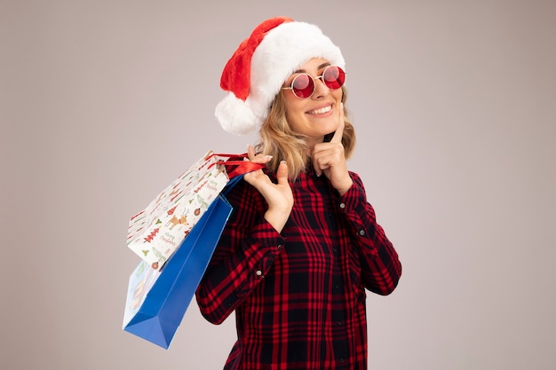 Lächelndes junges schönes Mädchen mit Weihnachtsmütze mit Brille, das Geschenktüte auf der Schulter hält und den Finger auf die Wange legt, isoliert auf weißem Hintergrund