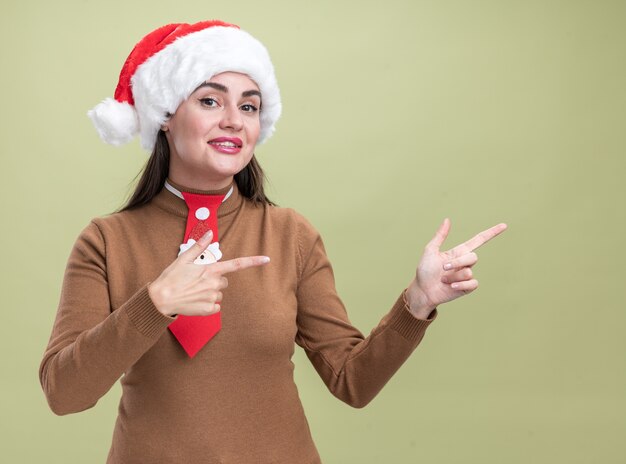 Lächelndes junges schönes mädchen mit weihnachtsmütze und krawattenpunkten an der seite isoliert auf olivgrüner wand mit kopierraum